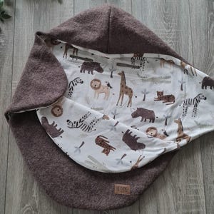 Peut inclure: Une couverture pour bébé marron avec une capuche. L'intérieur présente un tissu blanc imprimé d'animaux de safari, dont des lions, des girafes et des zèbres. La couverture porte une étiquette avec le mot "LIEBLING".