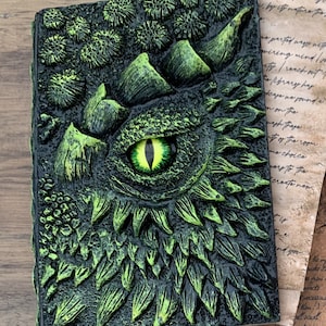 Puede incluir: Un diario encuadernado en cuero verde con un ojo de dragón y escamas en relieve en la portada.