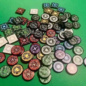 May include: A collection of small, flat game tokens in both square and circular shapes. The tokens feature various colours including red, green, blue, purple, gold, black, and white, with intricate geometric patterns and symbols printed on their surfaces.