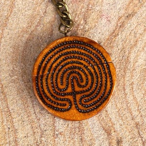 Può includere: Un portachiavi rotondo in pelle marrone chiaro con un disegno a labirinto nero. Il ciondolo è attaccato a una catena e a un portachiavi in metallo color bronzo. Un accessorio decorativo.