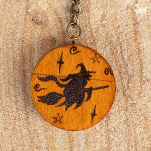 May include: A wooden Halloween keyring with a bronze-coloured ring and chain. The round wooden charm features a black silhouette of a witch on a broomstick, surrounded by stars and other symbols. The keyring is a decorative accessory.