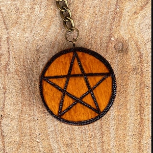 May include: A circular keyring with a bronze-coloured ring and chain. The pendant displays a pentagram design within a circle. The background is a light brown, textured surface.