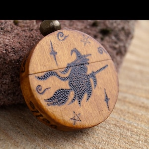 May include: A circular wooden pendant featuring a witch on a broomstick, etched in dark brown against a light brown background. The pendant has star and swirl details. It is attached to a brown cord, suitable for a necklace.