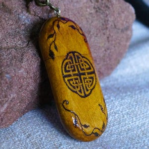 May include: An oval-shaped wooden pendant featuring a dark brown pyrography design of a central Celtic knot surrounded by delicate vine patterns. The pendant is finished with a glossy coating and is attached to a dark metallic chain link necklace.
