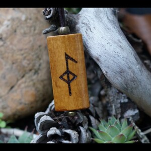 Peut inclure: Un pendentif en bois avec un cordon en cuir brun et un symbole de rune noir et blanc. Le pendentif est rectangulaire et a une finition en bois naturel.