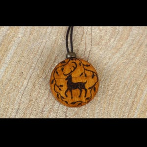May include: A round wooden pendant with a deer silhouette and leaf design. The deer is dark brown, and the pendant is attached to a black cord. The pendant is approximately 1.5 inches in diameter.