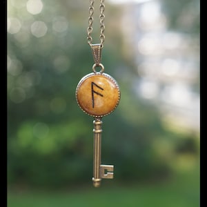Peut inclure: Un collier avec un pendentif en forme de clé en or et un charme rond en bois représentant la rune nordique Fehu. La rune est gravée à l'encre noire sur le charme en bois.