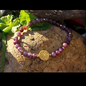 Peut inclure: Un bracelet de perles avec des pierres précieuses violettes profondes à facettes et de petites perles dorées. Le bracelet comprend un charm circulaire doré avec un motif d'arbre. Le bracelet est présenté sur une surface brune texturée.
