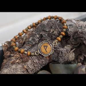 Peut inclure: Un bracelet en perles de bois brun avec un charme en métal représentant un symbole de rune. Le charme est de forme circulaire avec une bordure en forme de fleur.