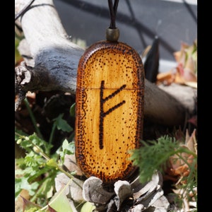 May include: A wooden pendant with a burned-in design of the Fehu rune, a symbol of prosperity and abundance in Norse mythology. The pendant is oval-shaped and has a brown leather cord.