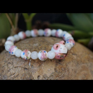 Op de afbeelding: Een kralenarmband met een keramische Maneki-neko kattenbedel. De armband heeft witte en bloemversierde kralen. De kattenbedel heeft een roze kop en gouden accenten. De armband ligt op een stenen ondergrond.