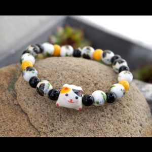 Op de afbeelding: Een kralenarmband met een keramische Maneki Neko kattenbedel. De armband bevat witte, zwarte en gele kralen met bloemen en abstracte patronen. De kattenbedel heeft een rood, zwart en oranje ontwerp.