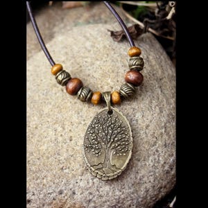 May include: A bronze tree of life pendant necklace. The oval pendant hangs from a brown leather cord, interspersed with wooden and metal beads. The necklace is displayed on a textured stone surface.