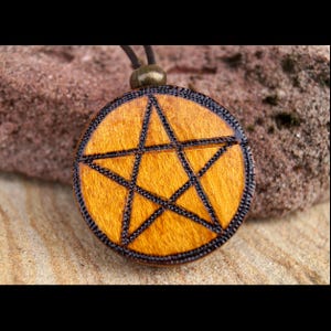 May include: A circular wooden pendant featuring a pentagram design. The pendant has a light brown centre with a black star outline and a black border. It hangs from a dark brown cord with a bronze-coloured clasp. The pendant rests on a sandy surface.