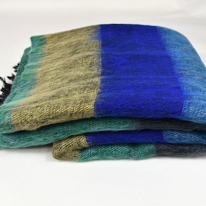 Op de afbeelding: Een gevouwen gestreepte deken met een blauw, groen en gouden kleurenschema. De deken heeft een franje rand.