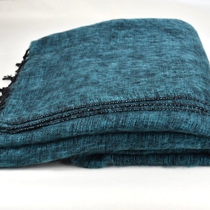 May include: Folded teal blanket with black fringe. The blanket has a textured appearance and is made of a soft material. The edges are finished with a dark thread. This blanket is perfect for home decor.