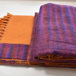 Puede incluir: Una manta suave de rayas naranja y morado con un borde de flecos. La manta está doblada por la mitad y el lado naranja está hacia arriba.
