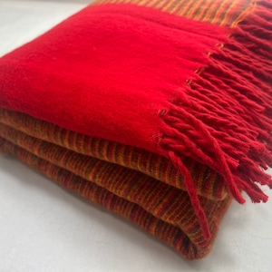 May include: A red and orange striped throw blanket with a red fringe. The blanket is folded in half and the fringe is visible on the bottom edge.