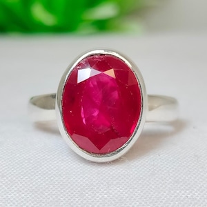 Peut inclure: Une bague en argent ornée d'un grand rubis de forme ovale. Le rubis est d'un rouge vif et est serti dans une monture simple en argent poli. La bague est présentée sur une surface blanche.