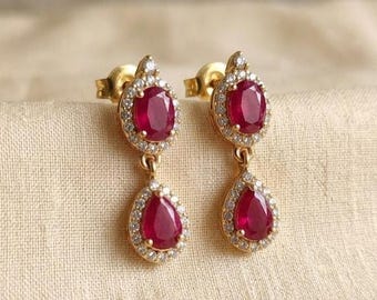 May include: A pair of gold drop earrings featuring oval-shaped red rubies at the top and pear-shaped red rubies at the bottom. Each ruby is surrounded by a halo of small white stones, with the two sections connected by a small gold link.