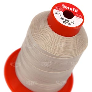 May include: A spool of beige sewing thread with a bright red plastic core. The top label reads Serafil, Amann Group, Col 0779, 30 Tex 90, and 900m. The thread is wound tightly around the cylindrical core, which features a wide red base and top.
