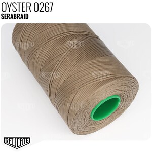 May include: A spool of Serabraid thread in a muted oyster color. The thread is tightly wound around a green plastic core. The text "OYSTER 0267" and "SERABRAID" are printed at the top of the image.