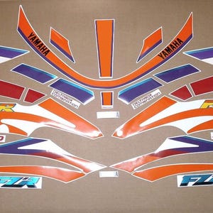 May include: A collection of orange, white, blue, and red Yamaha FZR motorcycle decals. The decals include the Yamaha logo, FZR, EXUP, and Genesis text. The decals are arranged on a brown background.