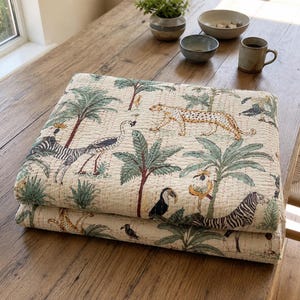 Puede incluir: Una colcha de algodón color crema plegada con un estampado repetitivo de temática selvática. El patrón incluye ilustraciones de leopardos, cebras, tucanes y loros intercalados entre palmeras verdes y diversas plantas tropicales.
