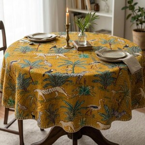 May include: A round mustard yellow tablecloth featuring a repeating pattern of green palm trees, white and black zebras, spotted cheetahs, and long-necked birds. The fabric has visible quilted stitching throughout and covers a circular wooden table.