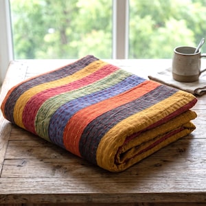 May include: A folded, striped quilt with a variety of colors including yellow, orange, red, green, and blue. The quilt is made of a soft material and is folded neatly on a wooden table. A mug and books are in the background.