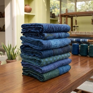 May include: A stack of folded rectangular quilts featuring various shades of blue and green fabric with visible running stitch patterns. The textiles are layered on a wooden surface, displaying a mix of solid colours and subtle geometric patterns.