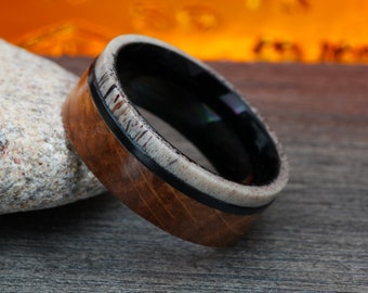 May include: A wooden ring with a black inlay and a white antler band. The ring is on a wooden surface.