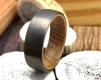 May include: A black tungsten carbide ring with a wood inlay. The ring is on a white background.