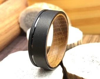 May include: A black tungsten carbide ring with a black inlay and a wood inlay. The ring is on a wooden surface.