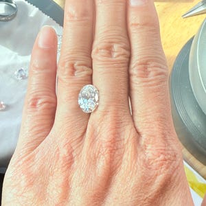 May include: A clear oval-shaped diamond gemstone on a finger.