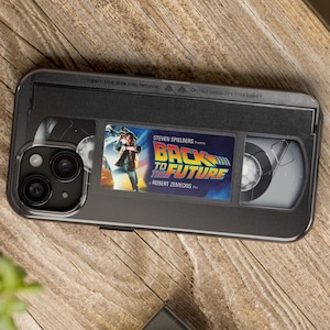 May include: A black phone case designed to resemble a VHS tape, with the "Back to the Future" movie poster. The case has a clear section for the phone's camera. It is set on a wooden surface.
