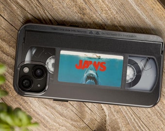May include: A black phone case with a VHS tape design featuring the Jaws movie poster. The case has a black and white image of a shark with the word "JAWS" in red letters. The case has a cutout for the phone's camera lens.