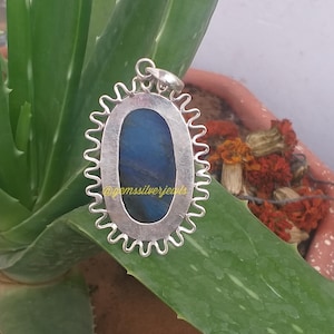 Op de afbeelding: Zilveren hanger met een blauwe edelsteen, gezet in een ovale lijst met een golvende rand. De hanger ligt op een groen alo&euml; vera blad. De tekst "@gemssilverjewels" is zichtbaar.