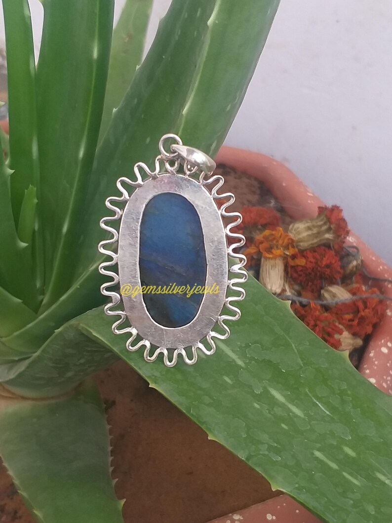 Op de afbeelding: Zilveren hanger met een blauwe edelsteen, gezet in een ovale lijst met een golvende rand. De hanger ligt op een groen alo&euml; vera blad. De tekst "@gemssilverjewels" is zichtbaar.