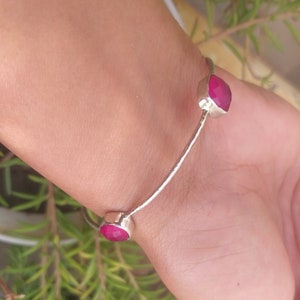 Könnte beinhalten: Ein silbernes Armreif-Armband mit zwei rosafarbenen Edelstein-Akzenten. Das Armband ist offen und hat ein schlichtes Design.