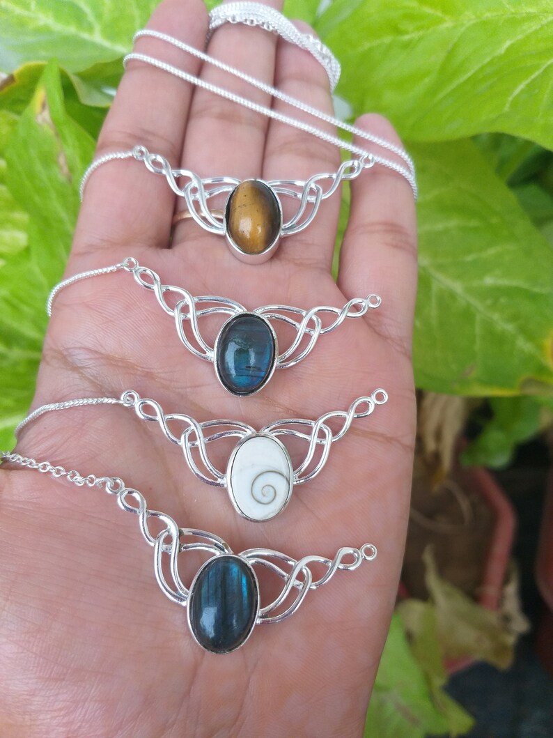 May include: Four silver necklaces with different oval-shaped gemstones. The necklaces feature a Celtic knot design. The gemstones are tiger's eye, labradorite, a white shell with a spiral pattern, and blue labradorite.