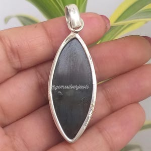 May include: A silver pendant with a dark, oblong stone. The stone is set in a silver frame and has a smooth, polished surface. The pendant is held by a hand, with the text "@gemssilverjewls" visible on the stone.