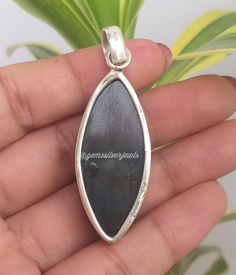 May include: A silver pendant with a dark, oblong stone. The stone is set in a silver frame and has a smooth, polished surface. The pendant is held by a hand, with the text "@gemssilverjewls" visible on the stone.