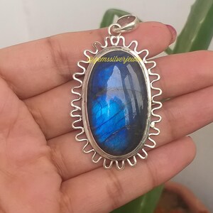 Op de afbeelding: Zilveren hanger met een ovale labradorietsteen. De steen vertoont iriserende blauwe en groene tinten. De hanger heeft een decoratieve, geschulpte rand. De tekst "@emssilverjew" is zichtbaar.