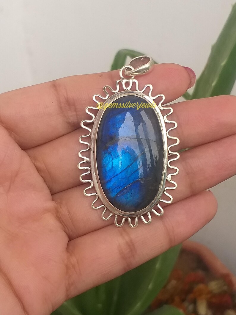 Op de afbeelding: Zilveren hanger met een ovale labradorietsteen. De steen vertoont iriserende blauwe en groene tinten. De hanger heeft een decoratieve, geschulpte rand. De tekst "@emssilverjew" is zichtbaar.