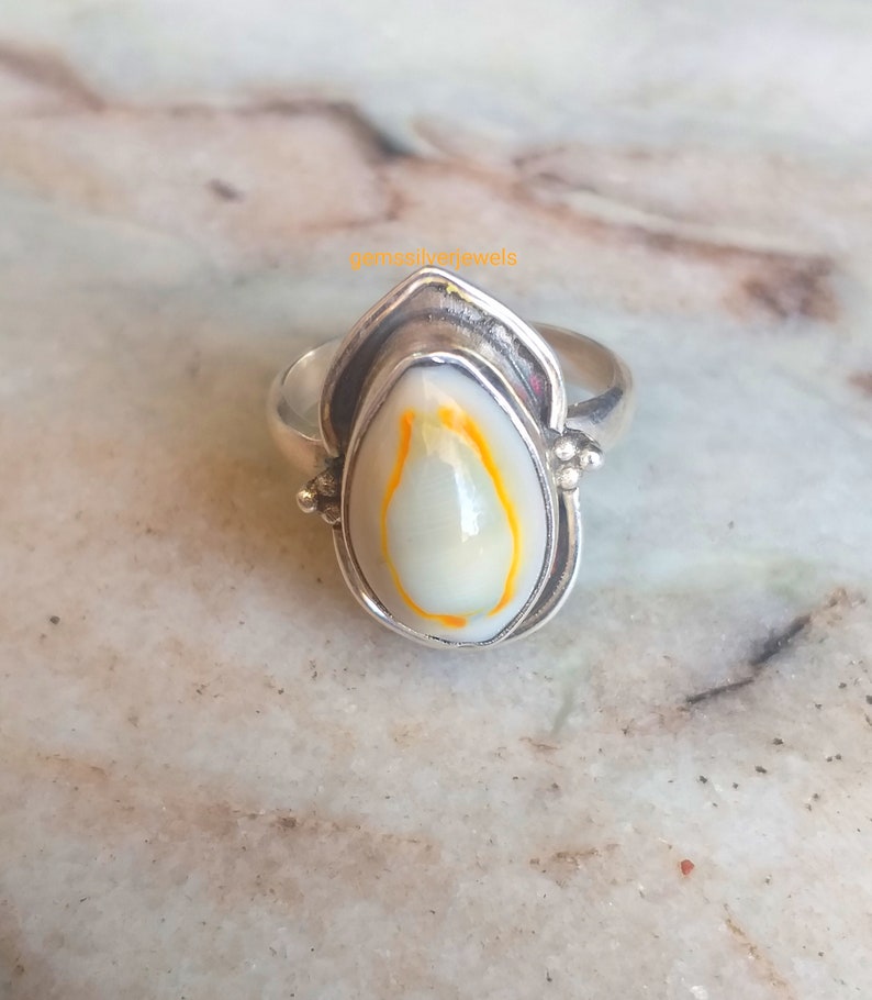 K&ouml;nnte beinhalten: Ein silberner Ring mit einem wei&szlig;en und orangefarbenen Muschelkamee, der in eine silberne Fassung gefasst ist. Der Ring liegt auf einer Marmorfl&auml;che.
