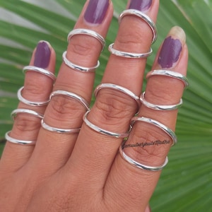 Peut inclure: Une main portant plusieurs bagues de doigt en argent. Les bagues sont de simples bandes arrondies qui encerclent les doigts à différents endroits. Les ongles sont peints avec du vernis à ongles violet. En arrière-plan, une feuille verte.