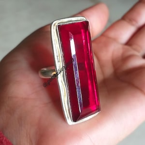 Peut inclure: Bague en argent ornée d'une grande pierre précieuse rouge rubis rectangulaire. La pierre est sertie dans un cadre en argent. La bague est tenue dans une main. Le texte "@gemsilverjewels" est visible sur la bague.