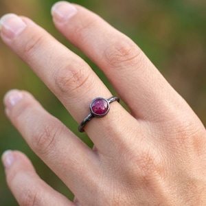 Puede incluir: Un anillo con una pequeña piedra preciosa redonda de color rosa intenso engastada en una banda de metal oscuro y texturizado. La banda tiene un acabado de superficie rugoso e irregular, y la piedra está en un engaste circular simple.