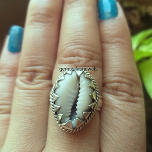 May include: A silver ring with a white cowrie shell set in a silver bezel. The shell is surrounded by a silver rope design and a row of silver triangles.